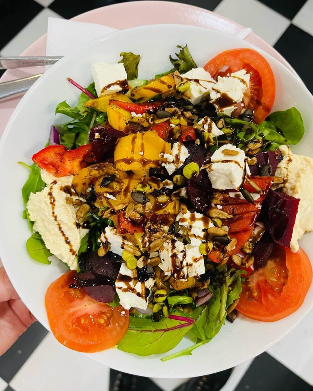 A colorful salad with greens, tomatoes, feta, beets, seeds, hummus, and drizzled with balsamic vinaigrette in a white bowl. - Home Instead