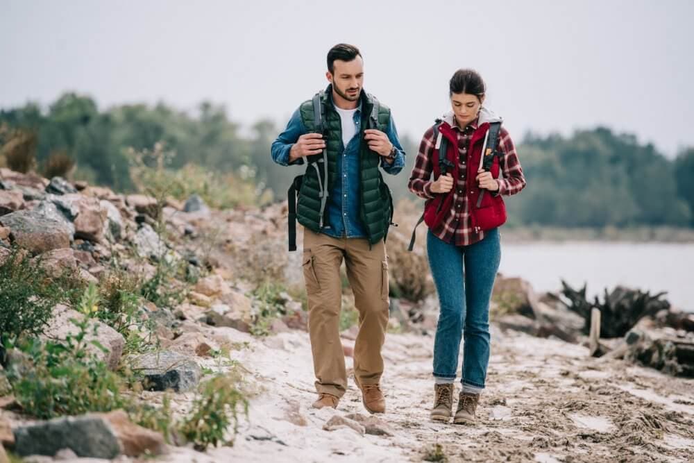 A man and a woman, wearing backpacks and outdoor gear, walk along a rocky lakeside path. - Home Instead