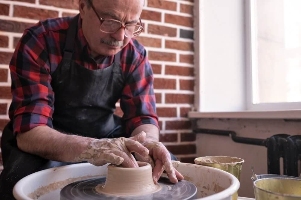 Man throwing clay on a pottery wheel in a studio with brick walls and window light. - Home Instead