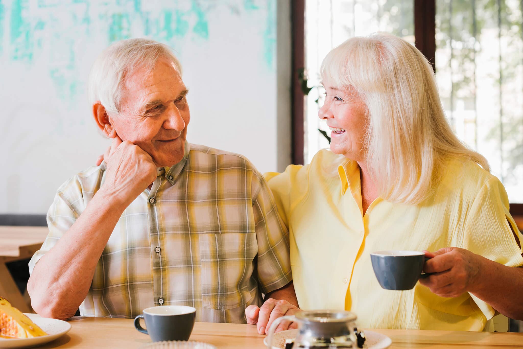 Elderly couple drinking coffee and smiling at each other while sitting at a table. - Home Instead
