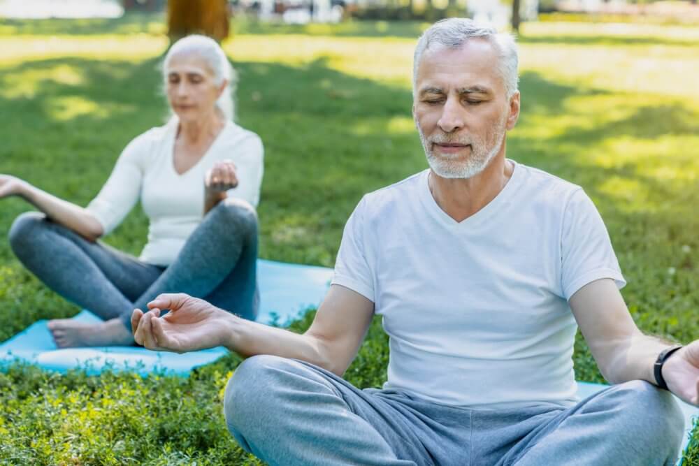 Two elderly individuals meditate in a park, sitting cross-legged on the grass with eyes closed and hands in mudra. - Home Instead