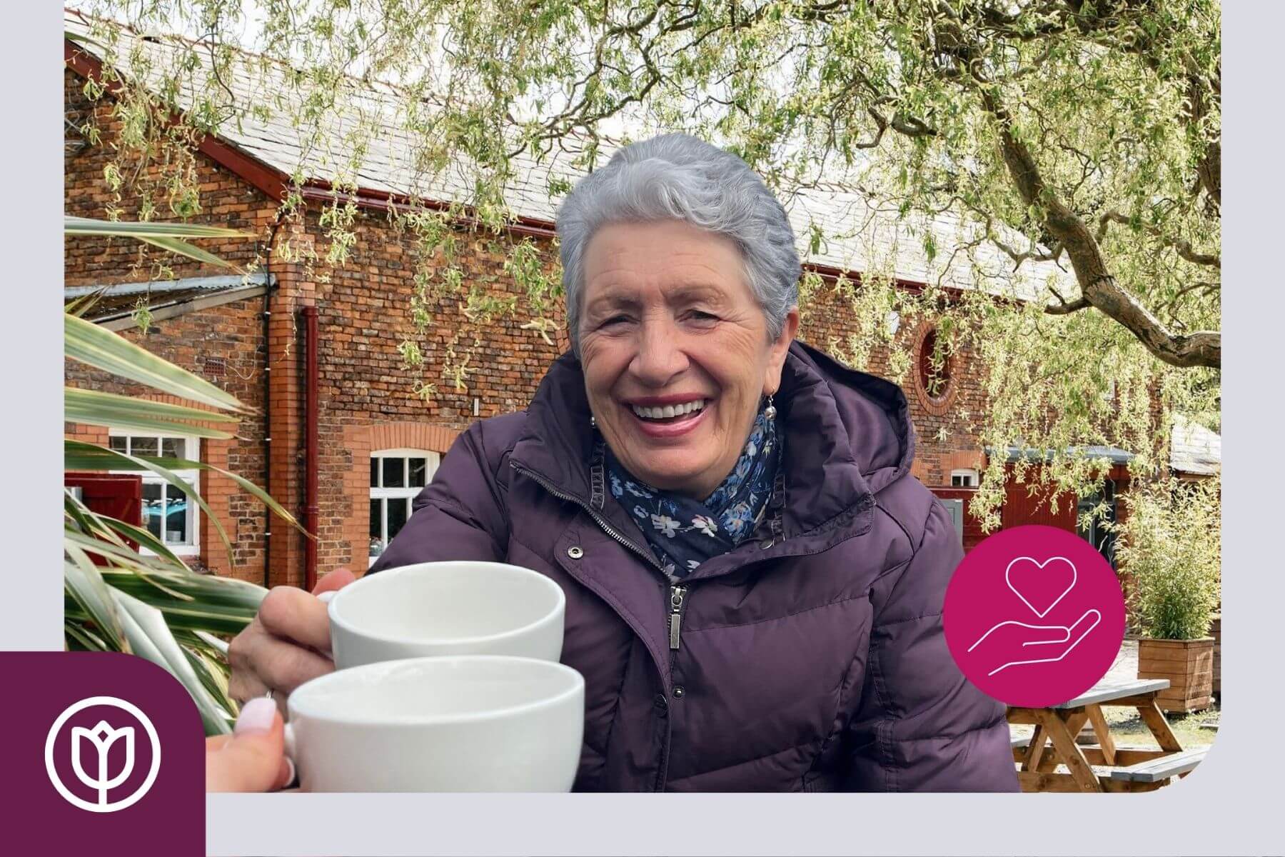 An elderly woman in a purple jacket smiles while toasting cups outdoors; a heart in hand icon is in the lower right. - Home Instead