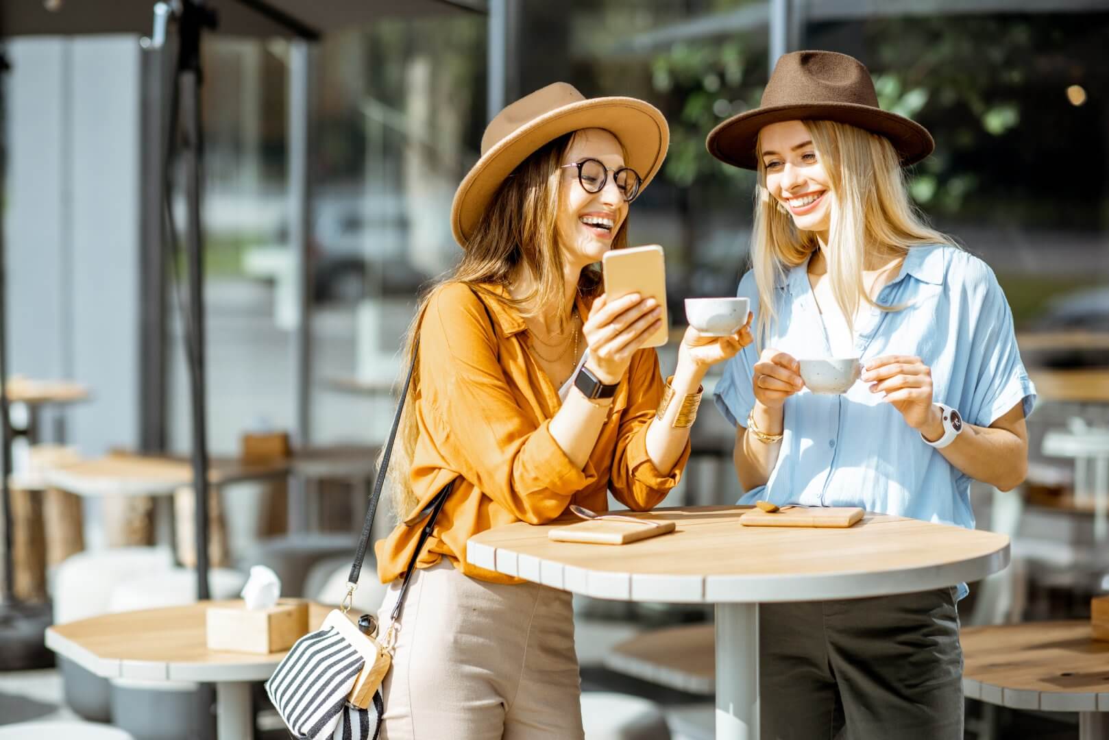 Two women drinking coffee and laughing, one holding a phone, both wearing hats at an outdoor café table. - Home Instead