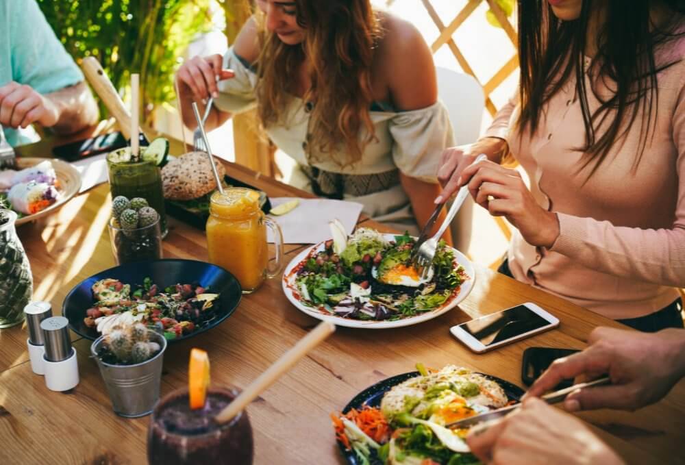 People enjoying a meal with various dishes and drinks on a wooden table outdoors. - Home Instead
