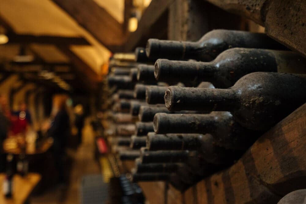 Dusty wine bottles stored horizontally in a dimly lit wine cellar with wooden beams. - Home Instead