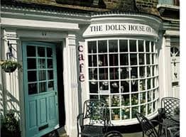 A charming café with a curved window, named "The Doll's House on the...", with an outdoor seating area and a blue door. - Home Instead