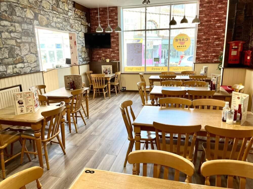 A cozy café with wooden tables and chairs, brick walls, a TV, and large windows letting in natural light. - Home Instead