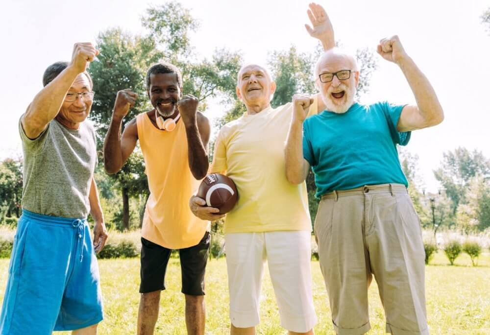 Five elderly men smile and fist pump in a park, one holding a football. The sun shines brightly in the background. - Home Instead