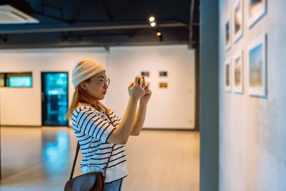 Person in a striped shirt and beanie taking a photo of artwork in a gallery setting. - Home Instead
