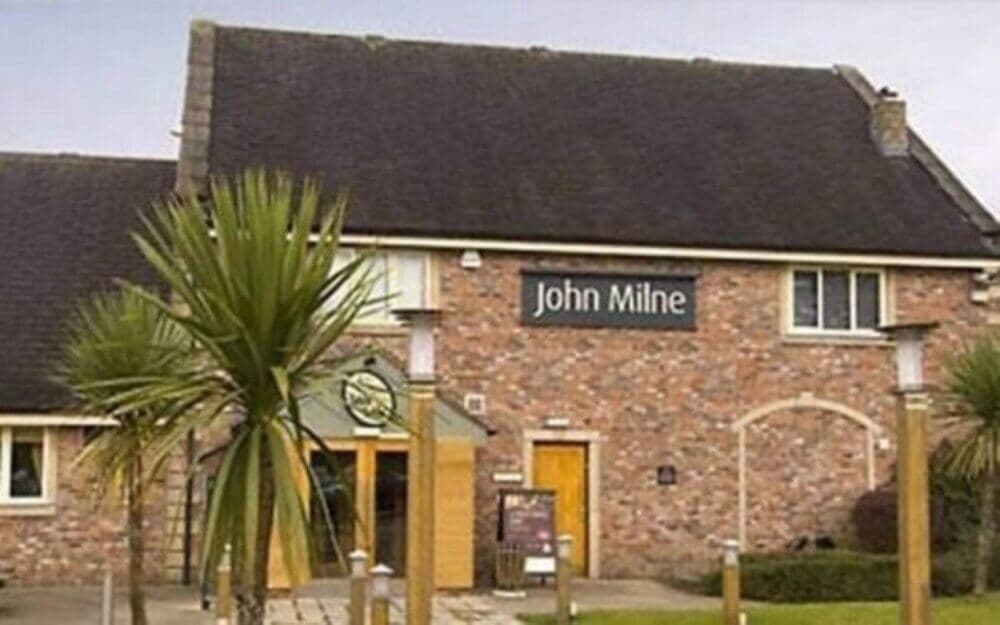 Exterior of a brick building with a sign reading "John Milne" above the entrance, surrounded by greenery and tall palms. - Home Instead