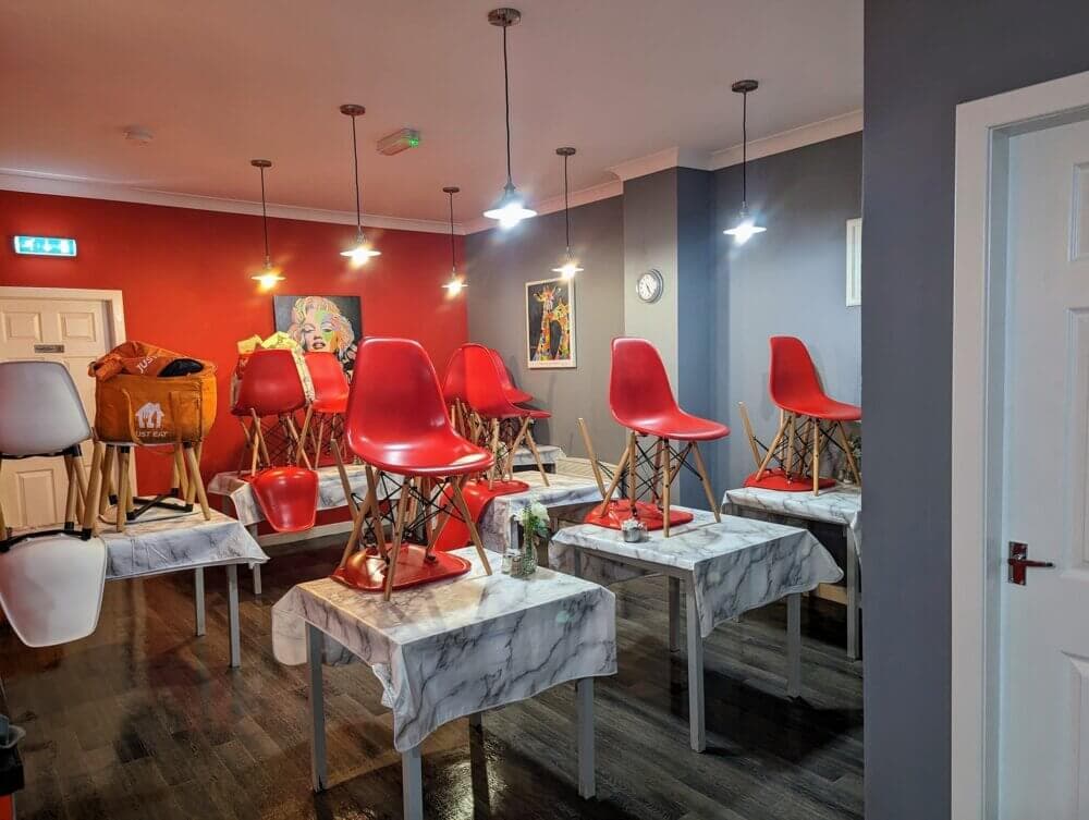 Dining room with red chairs stacked on tables, bright artwork on walls, and modern lighting fixtures hanging from ceiling. - Home Instead