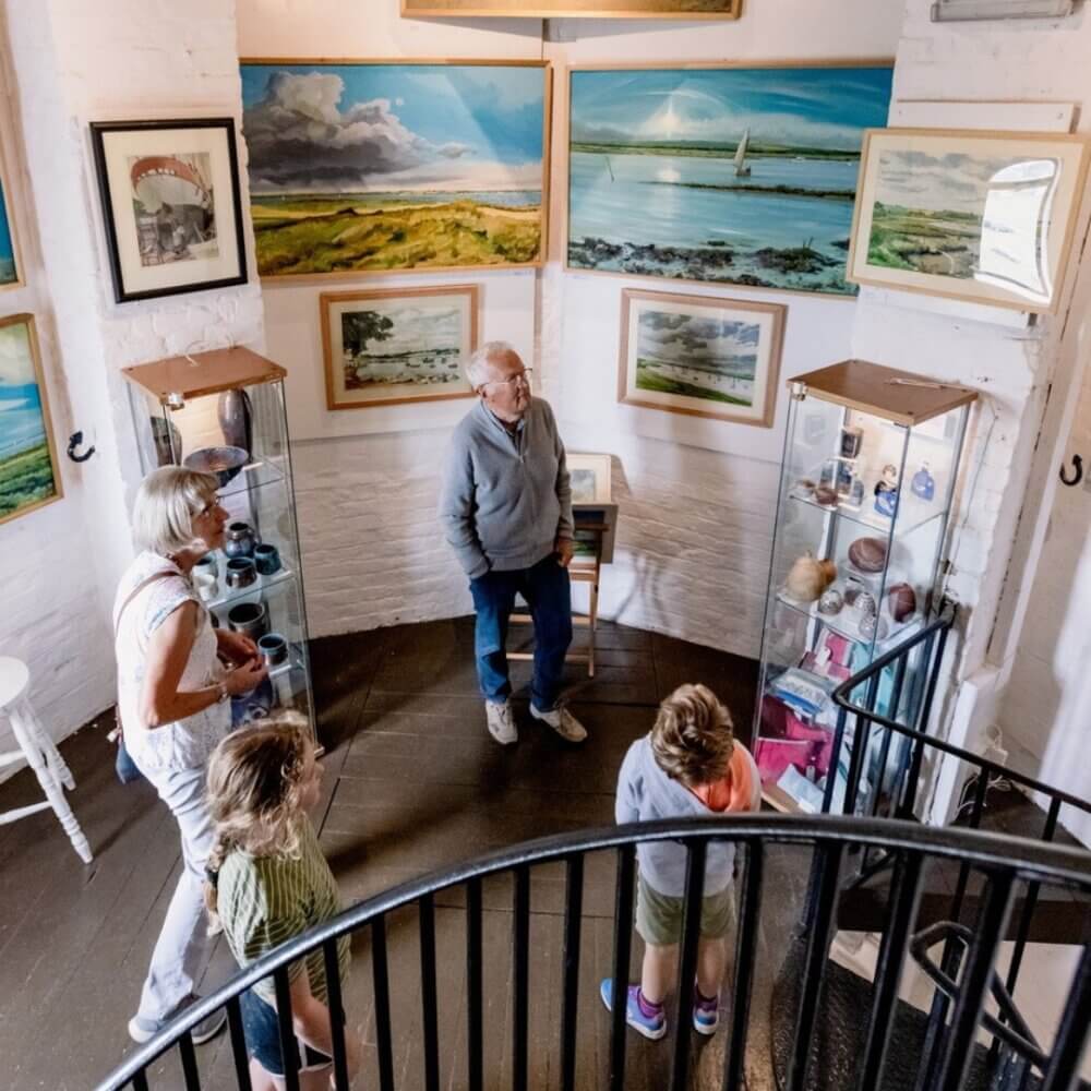 A group of people in a small art gallery observing paintings and ceramics displayed on the walls and in glass cases. - Home Instead