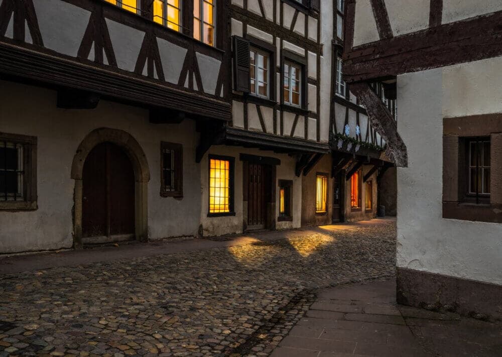 A cobblestone alley with lit windows in timber-framed buildings at dusk. - Home Instead