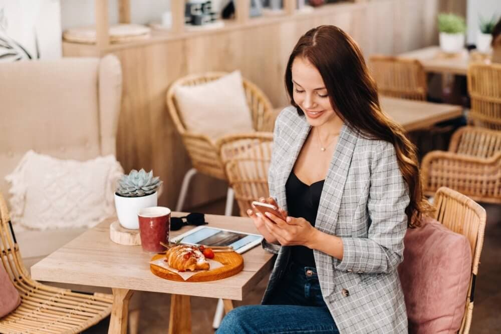 A woman in a plaid blazer is seated at a café table with pastries, coffee, a tablet, and a plant, smiling at her phone. - Home Instead