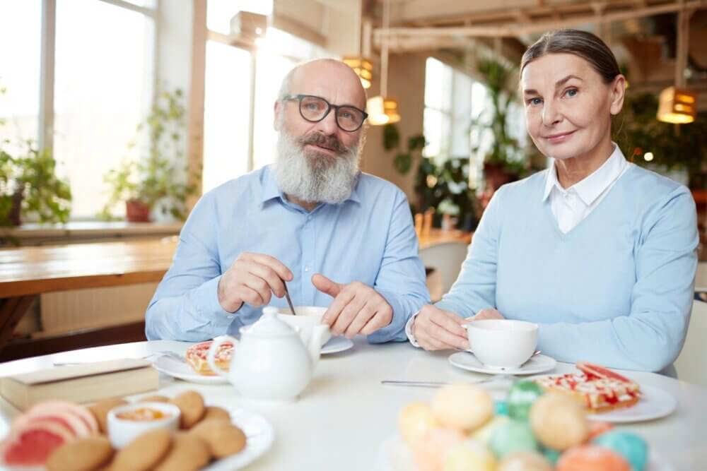 Two elderly people enjoying tea and pastries in a cozy cafe with a warm ambiance. - Home Instead