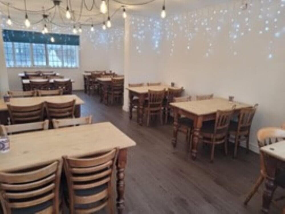 A cozy, empty restaurant with wooden tables and chairs, string lights on the ceiling, and fairy lights on the wall. - Home Instead