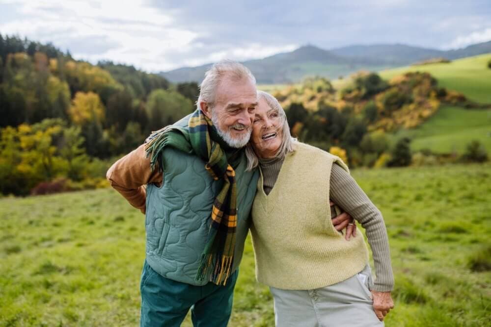 Elderly couple smiling and embracing in a lush, green outdoor setting with hills and trees in the background. - Home Instead