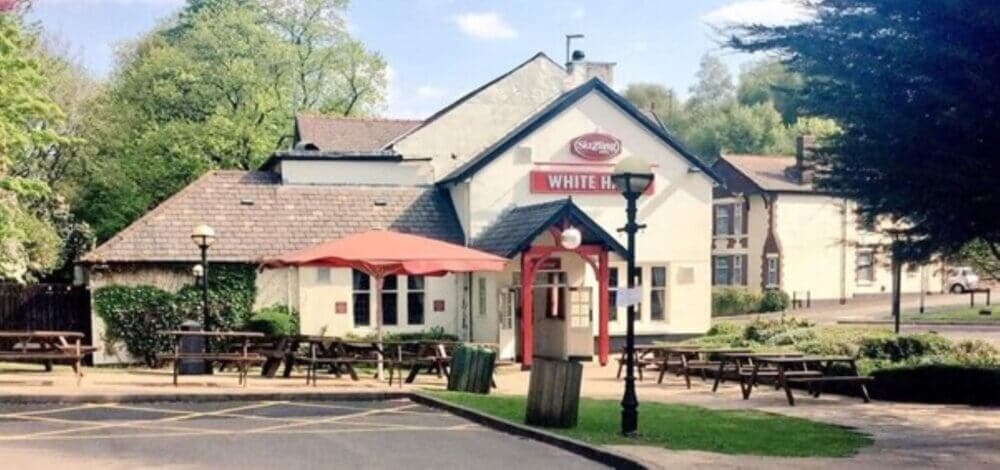 The outside area of a pub with red umbrellas, wooden picnic tables, and a sign reading "White Hart. - Home Instead