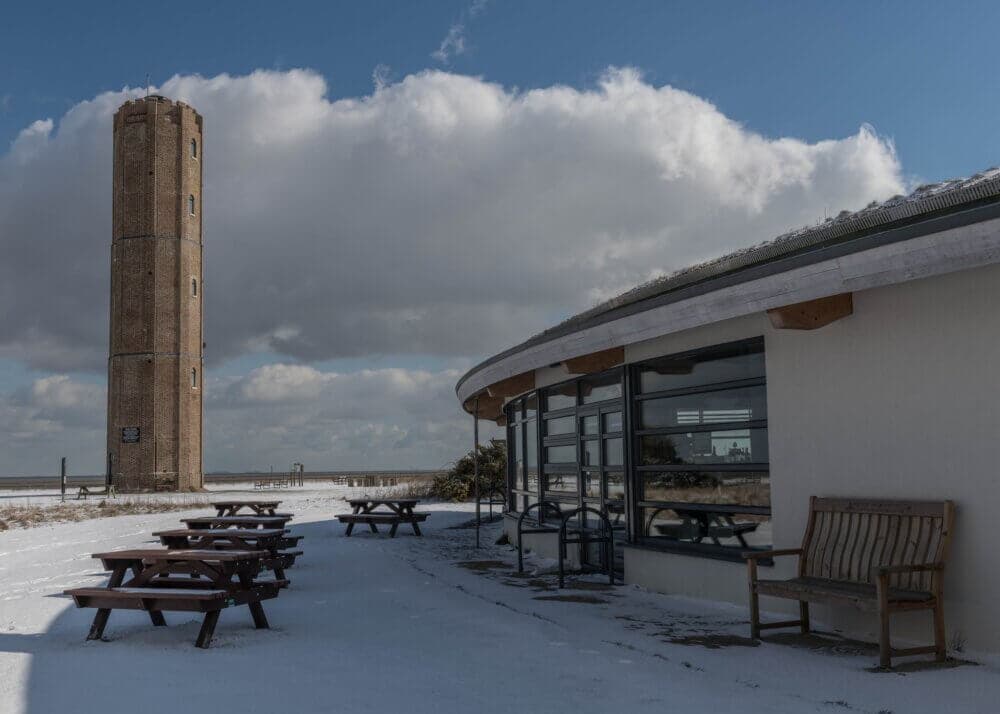 A snowy scene with an old brick tower, picnic tables, benches, and a building under a partly cloudy sky. - Home Instead
