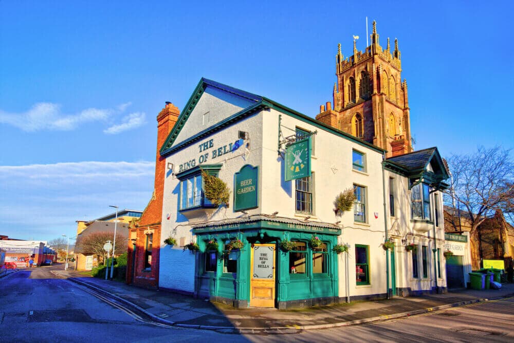 Image of "The Ring of Bells" pub with a church tower in the background, brightly lit by the sun. - Home Instead