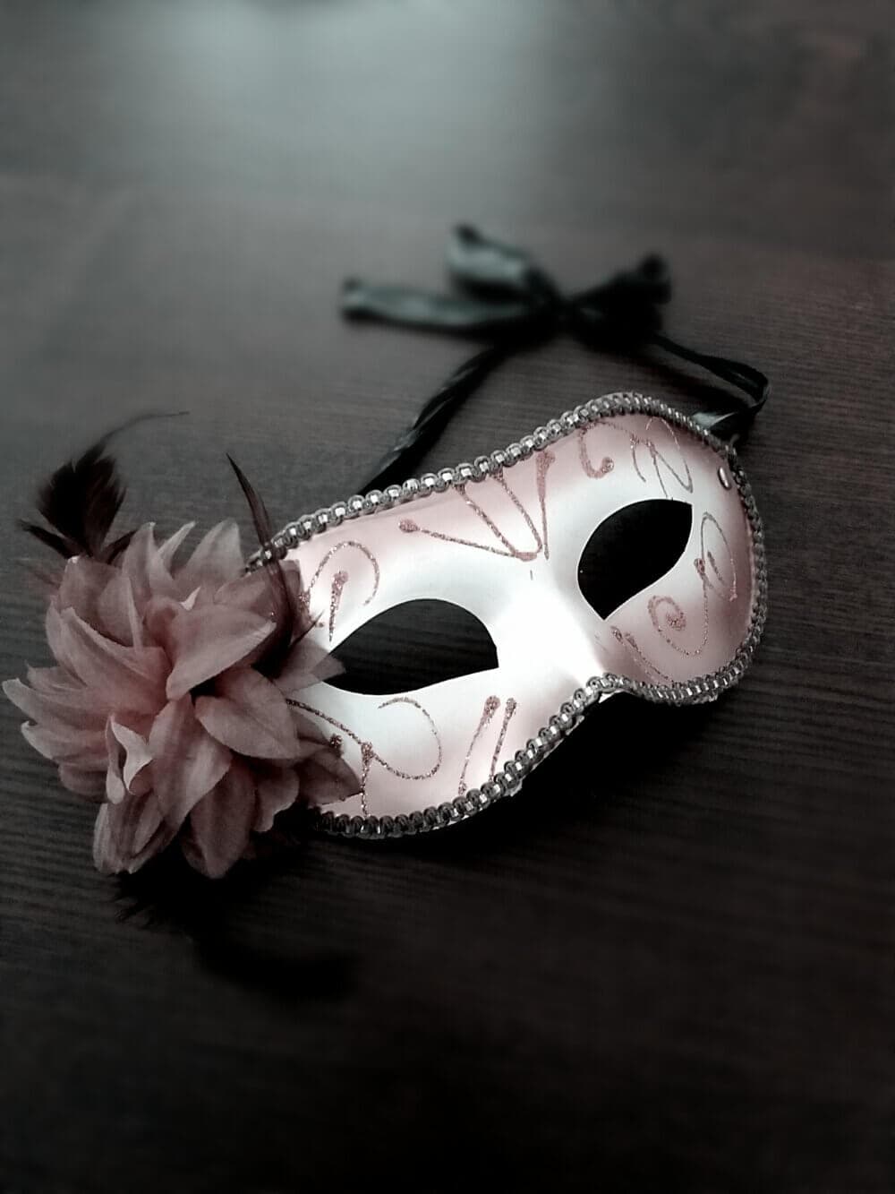 A decorative mask with feathers and a flower on one side, resting on a dark surface. - Home Instead