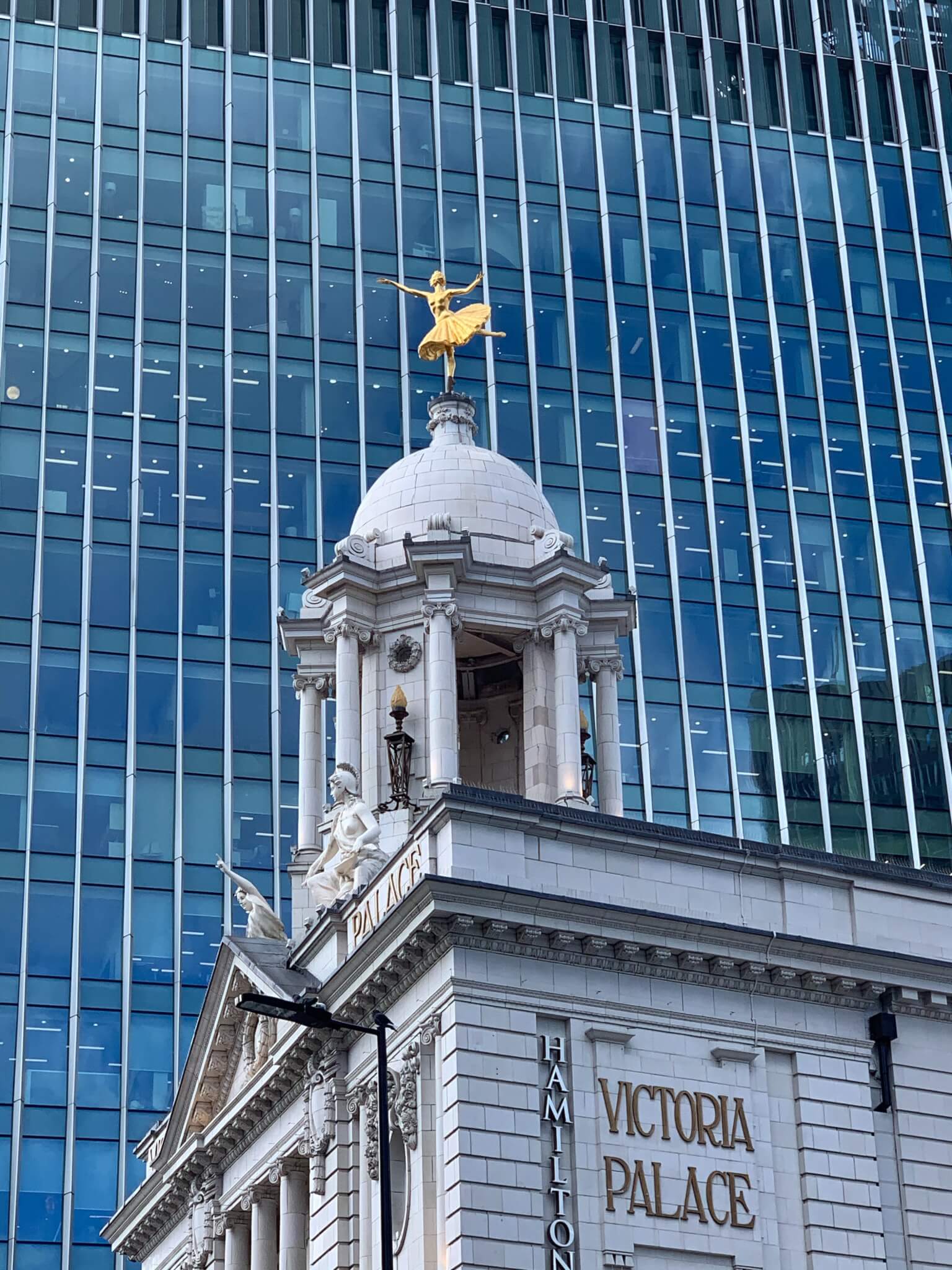 Victoria Palace Theatre with its golden statue against a backdrop of a modern skyscraper with glass windows. - Home Instead