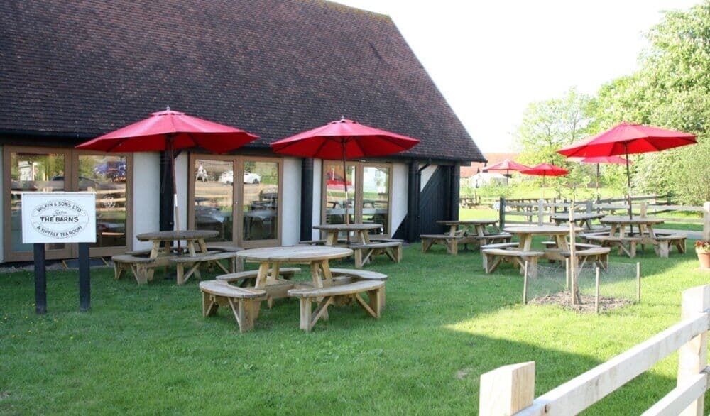 Outdoor seating area with wooden picnic tables, red umbrellas, and a sign reading "The Cressing Barns". Green grass and trees. Historic site Essex- Home Instead