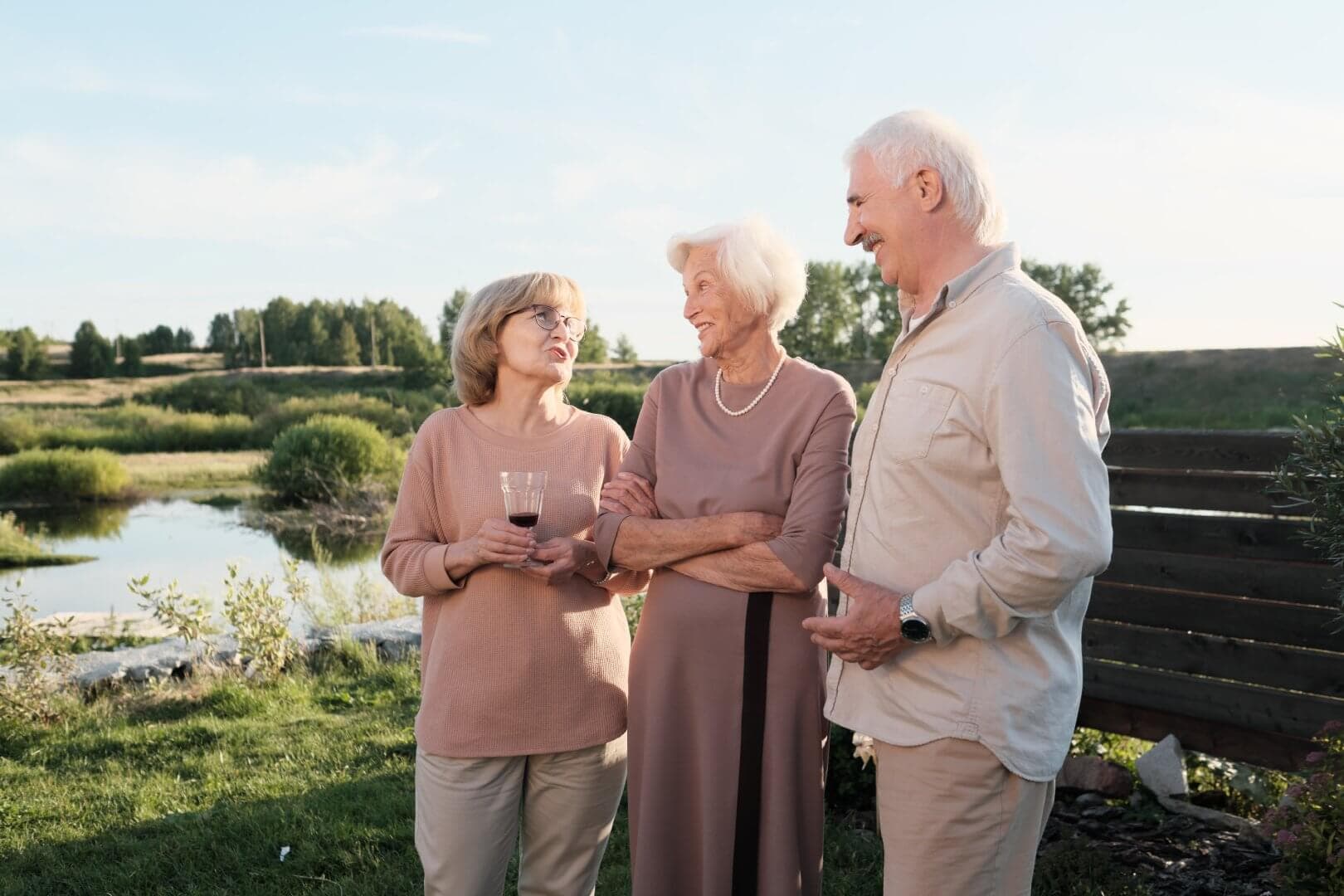 Three older adults stand outdoors by a pond, talking and smiling together on a sunny day. - Home Instead