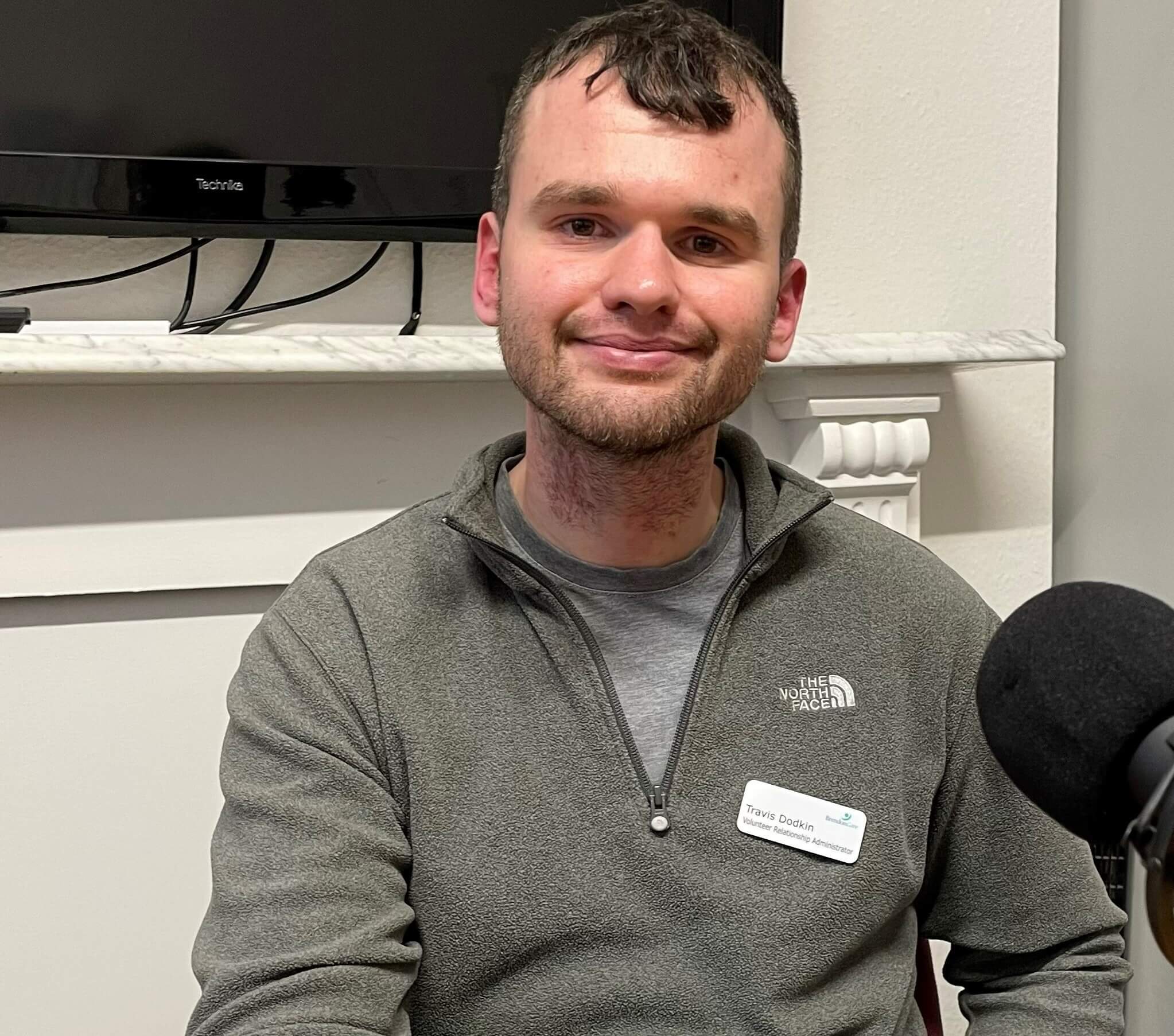 A man with a beard and short hair smiling while sitting. He wears a grey North Face jacket and a name tag. - Home Instead