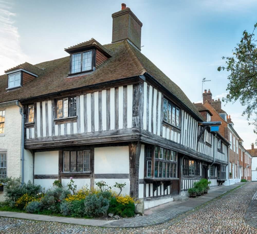 Medieval timber-framed house on a cobblestone street with a garden in front, under a blue sky. - Home Instead Southampton
