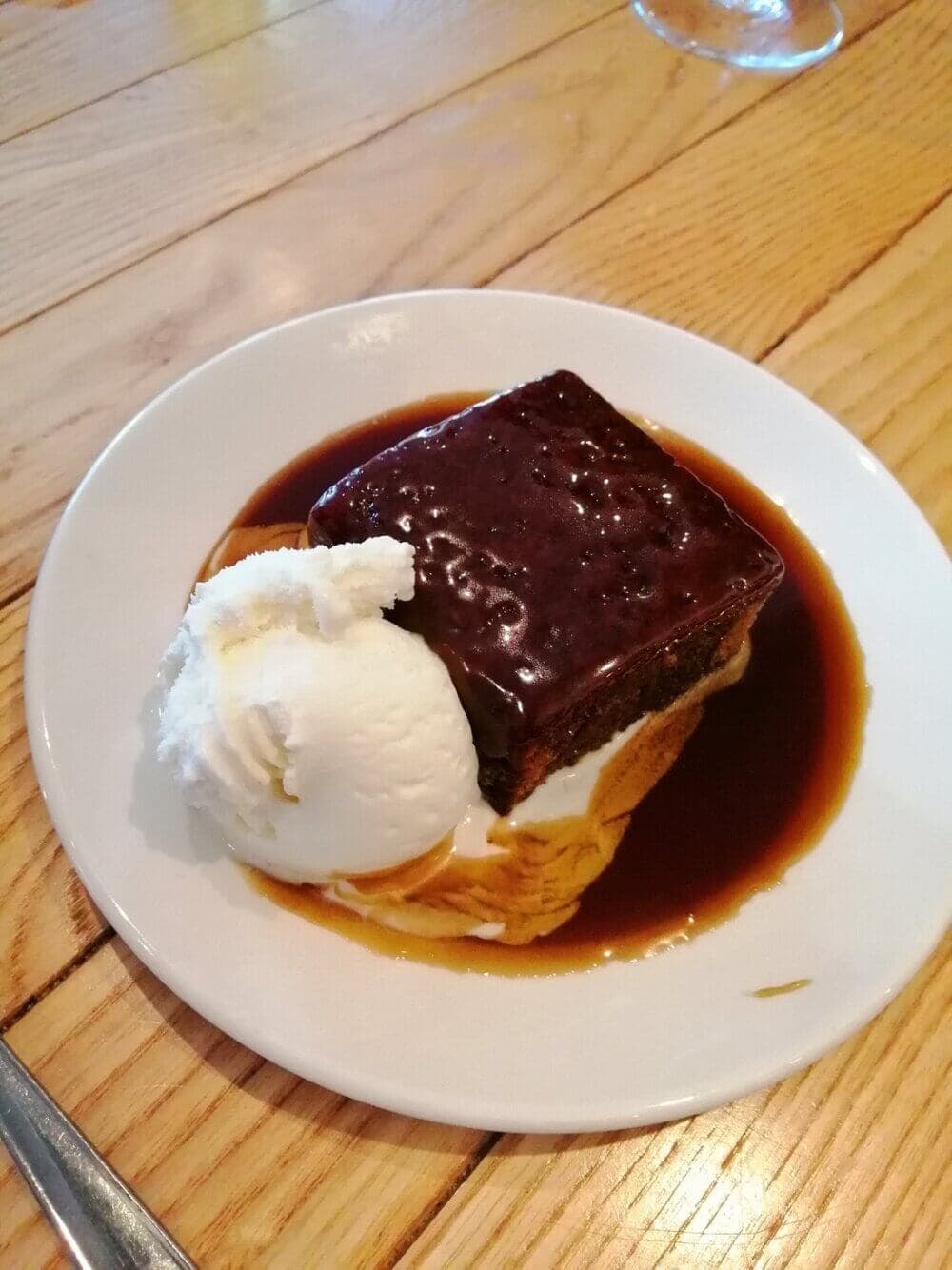 A plate with sticky toffee pudding, a scoop of vanilla ice cream, and a caramel sauce on a wooden table. - Home Instead