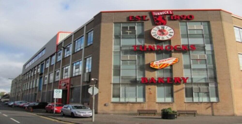A large brick building with "Tunnocks Bakery" signage, featuring a clock and a caramel wafer wrapper decoration. - Home Instead