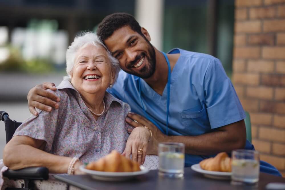 A smiling elderly woman and a young man in scrubs share a hug while sitting at an outdoor table with food and drinks. - Home Instead