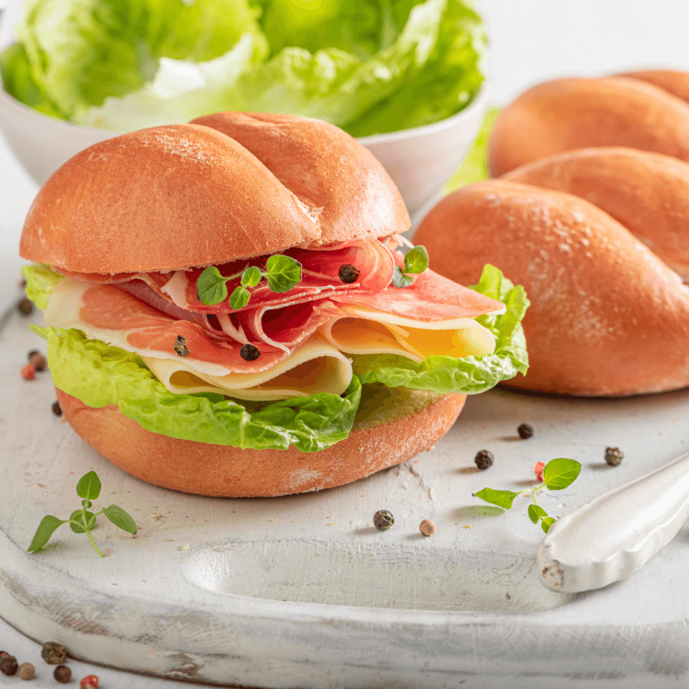 A sandwich with lettuce, cheese, and deli meat on a cutting board, accompanied by a bowl of lettuce and extra rolls. - Home Instead