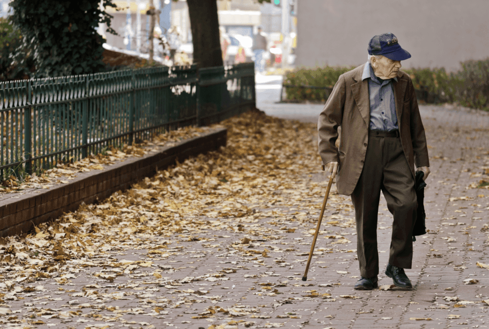 Elderly man in a brown suit and blue cap walking with a cane on a leaf-strewn sidewalk. - Home Instead