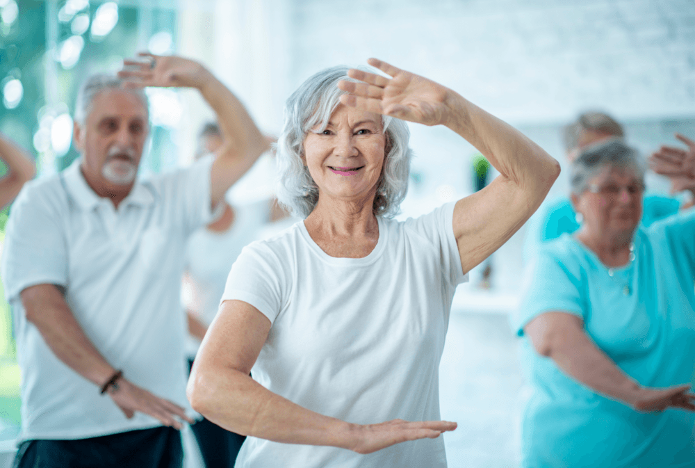 A group of seniors wearing white and blue shirts practice tai chi in a bright room, smiling and raising their hands gracefully. - Home Instead