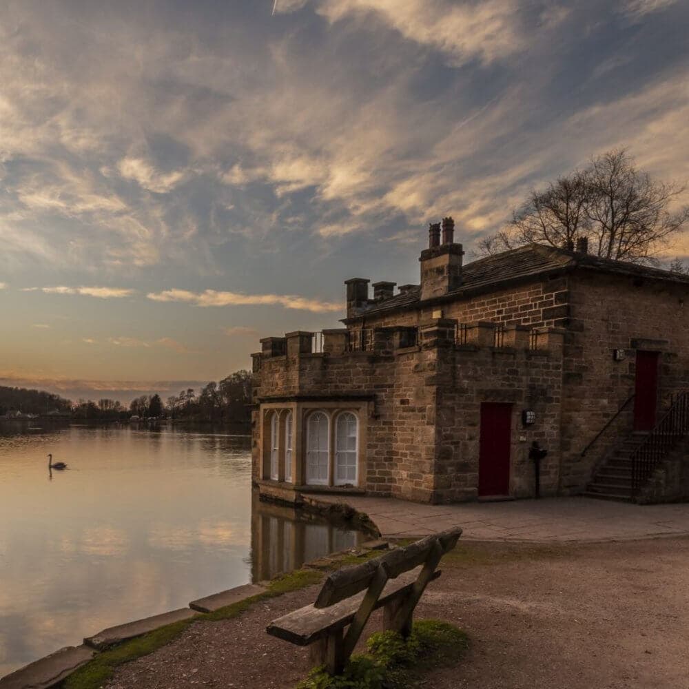 A stone building by a calm lake with a swan swimming nearby, under a cloudy sky at sunset. A wooden bench is in the foreground. - Home Instead