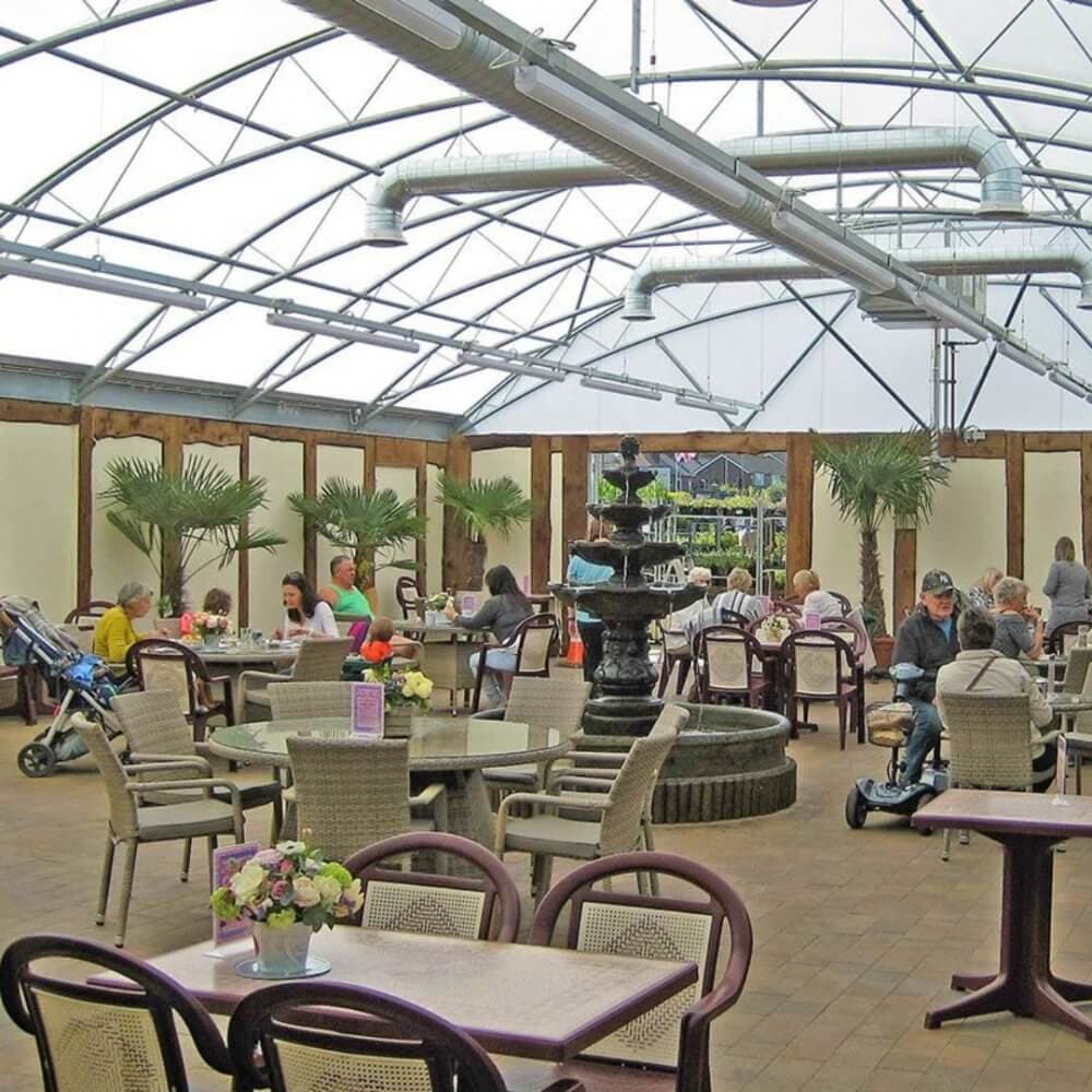 A bustling indoor café with tables, potted plants, a water fountain, and patrons enjoying their meals under a transparent roof. - Home Instead