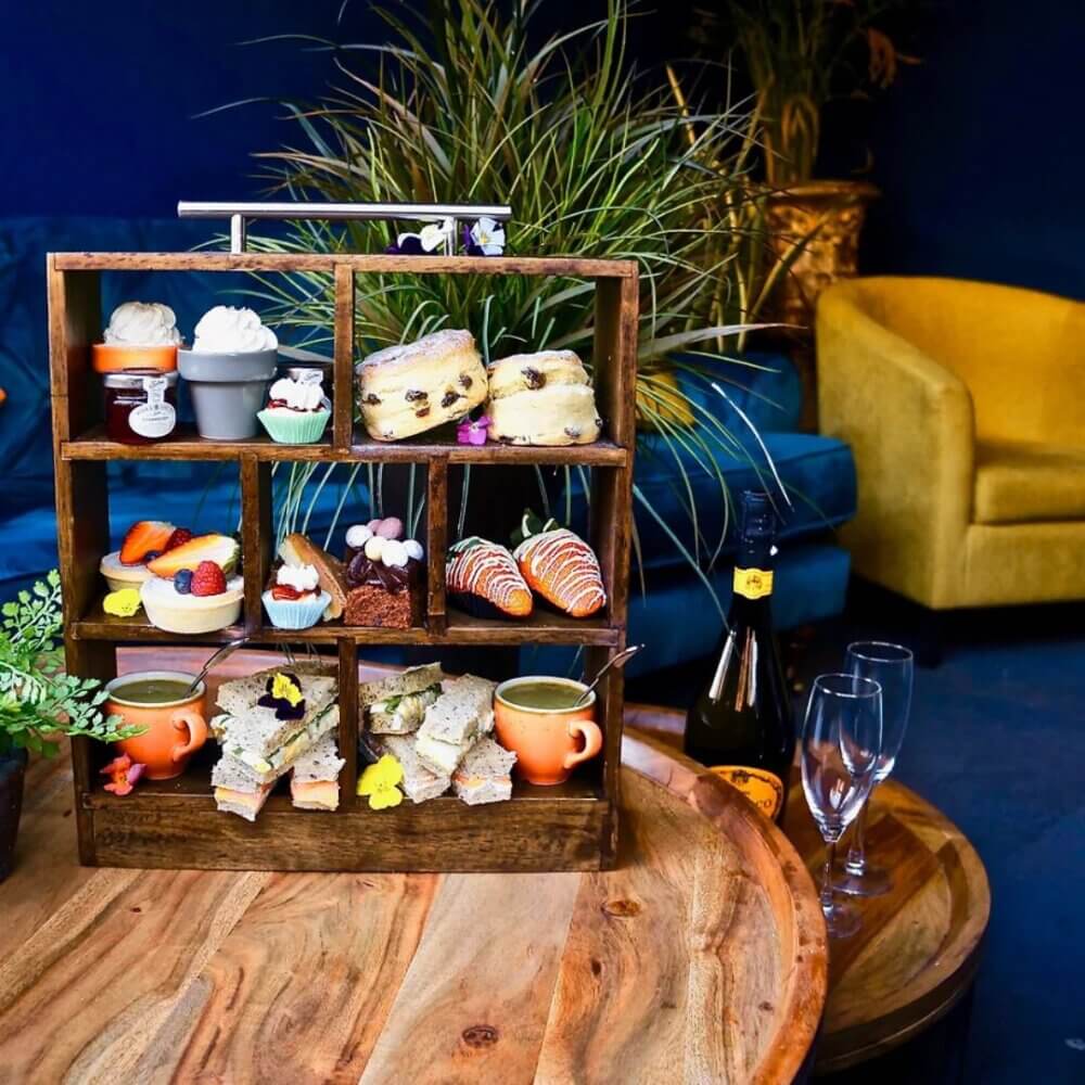 Tiered afternoon tea display with sandwiches, scones, and desserts on a wooden rack. Champagne bottle and glasses nearby. - Home Instead