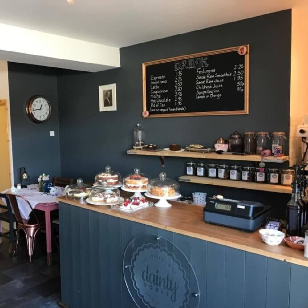 Cozy cafe with cakes on a counter, menu board on the wall, and tea jars on a shelf. - Home Instead