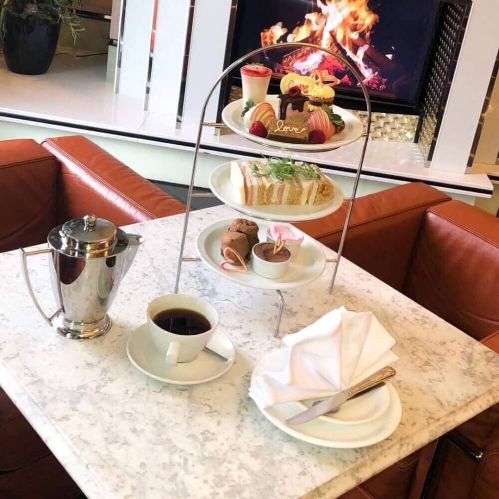 A table with tea, a three-tiered dessert stand, and a fireplace in the background. - Home Instead