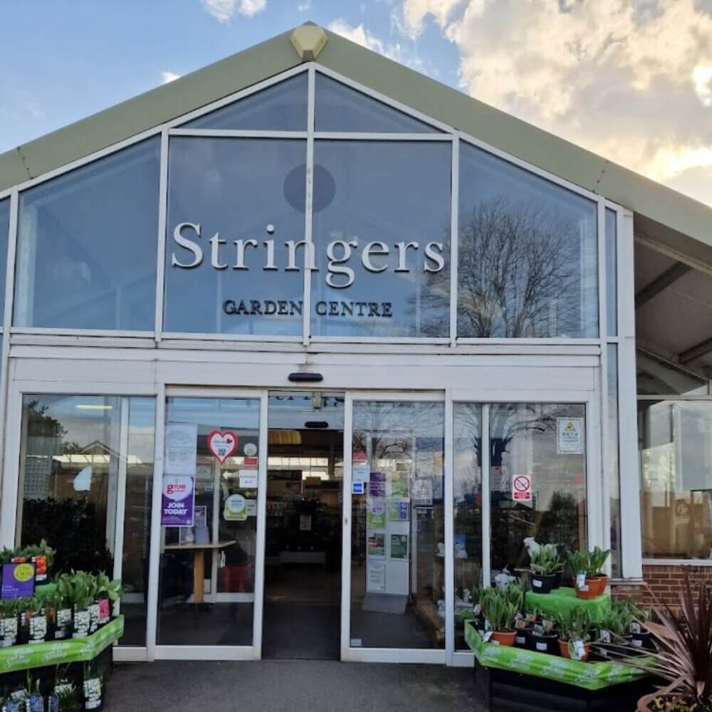 Front entrance of Stringers Garden Centre with plants on display outside and multiple signs on the glass doors. - Home Instead
