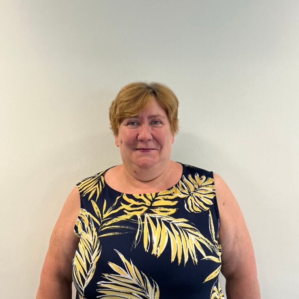 A woman with short hair in a navy blue and yellow leaf-patterned top stands against a plain white background. - Home Instead
