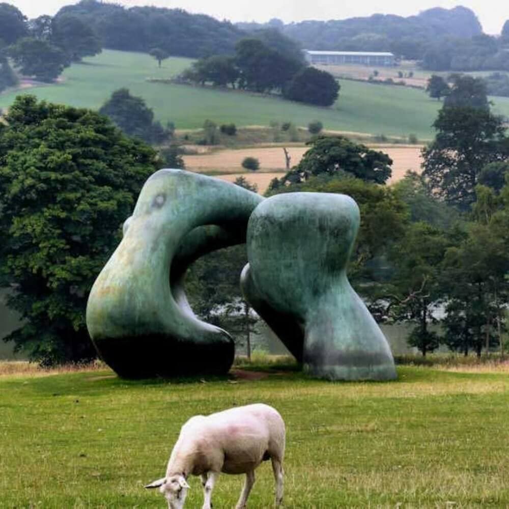 A sheep grazes in front of a large abstract sculpture on a grassy field with trees and hills in the background. - Home Instead