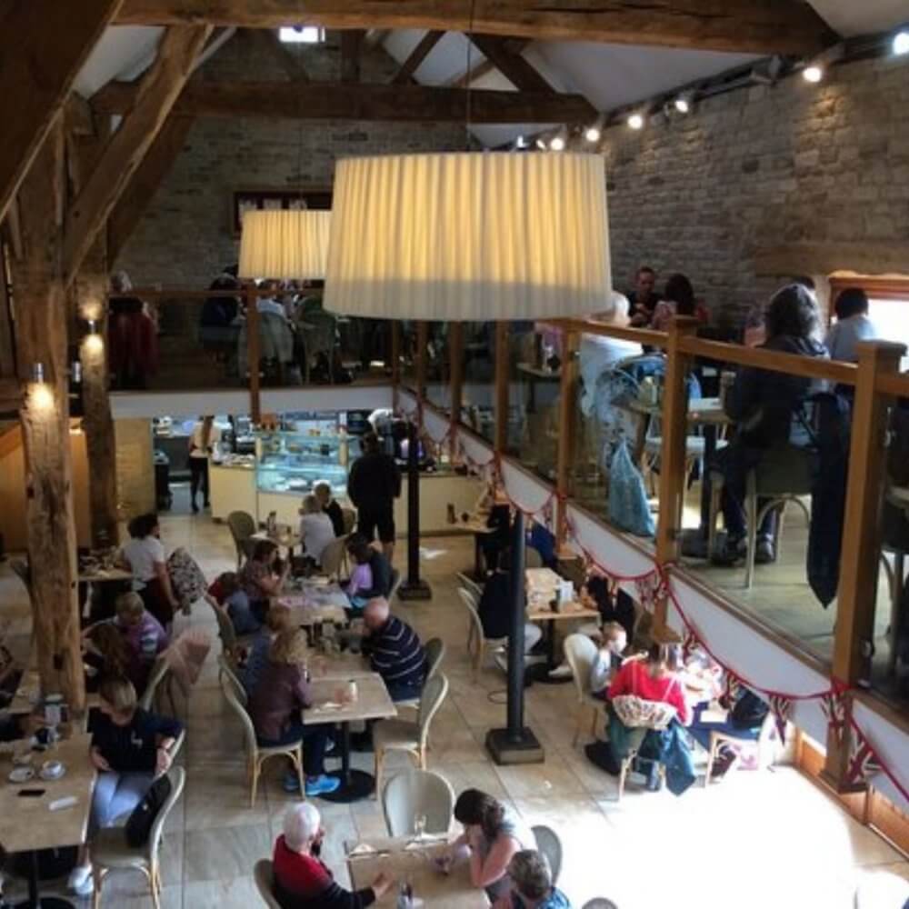 A busy, rustic café with wooden beams, two levels, and patrons enjoying their meals at tables. Large hanging lights. - Home Instead