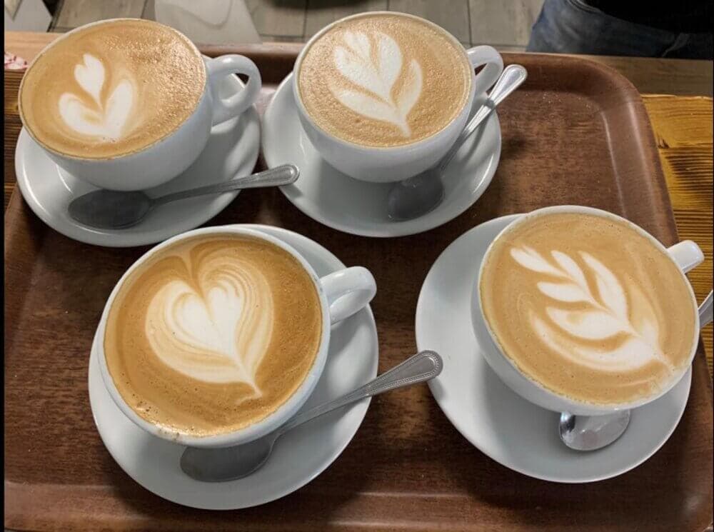 Four cups of latte with leaf-patterned foam art, arranged on a wooden tray with spoons beside each cup. - Home Instead