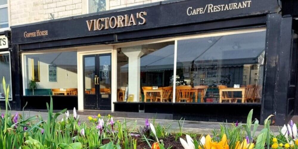 Front view of Victoria's Cafe/Restaurant with flowers in the foreground and wooden chairs and tables inside. - Home Instead