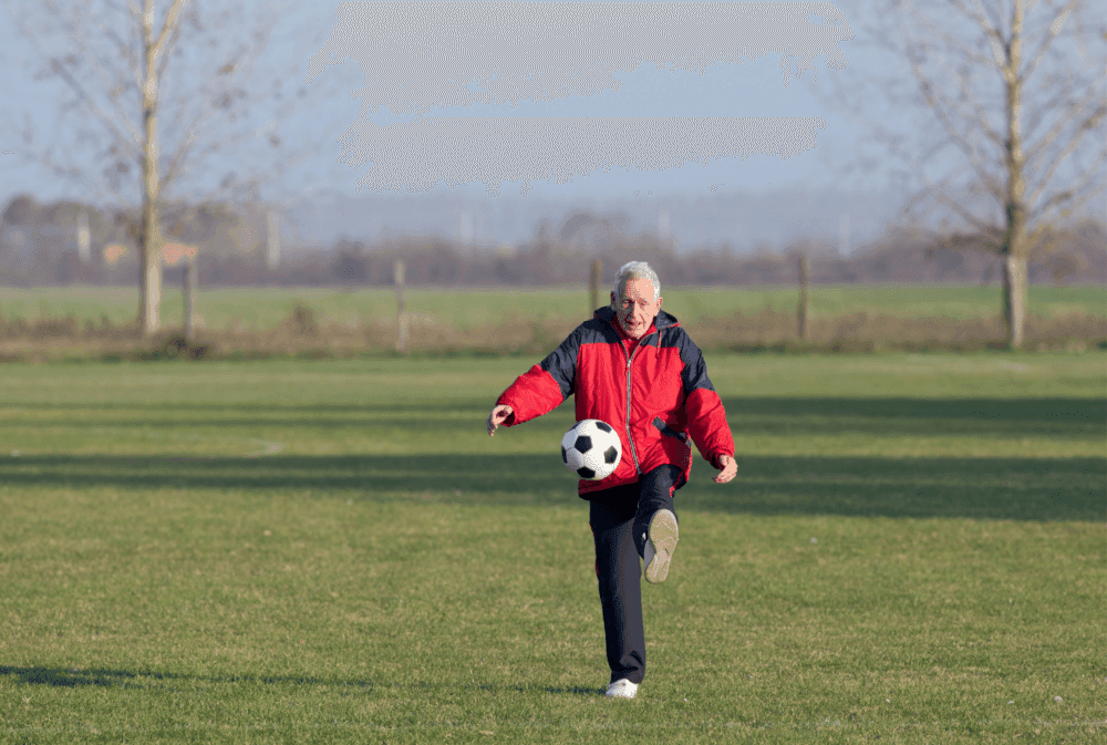 An elderly man in a red jacket kicks a soccer ball on a grassy field with trees and a fence in the background. - Home Instead
