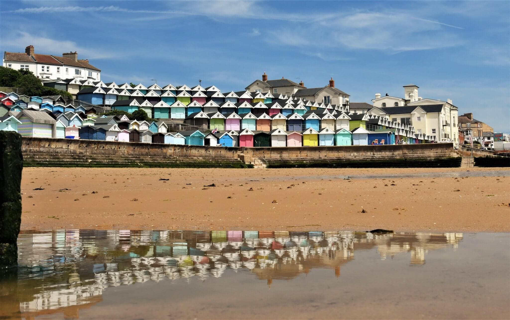 Colorful beach huts lined up on a seaside promenade, with their reflections visible in a shallow water puddle. - Home Instead