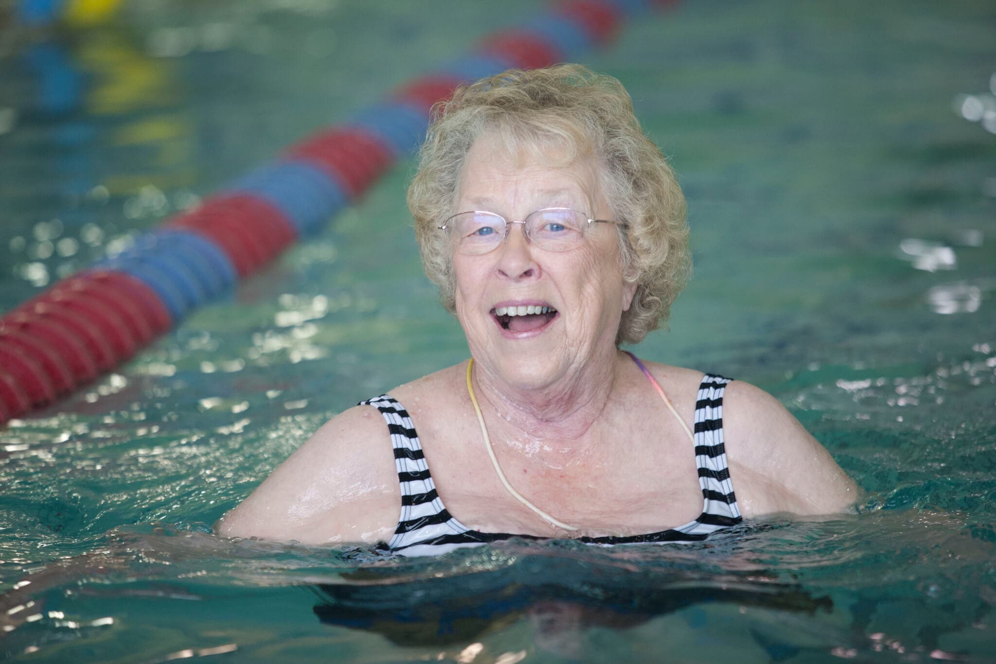 An older woman with glasses is smiling while swimming in a pool, wearing a black and white striped swimsuit. - Home Instead